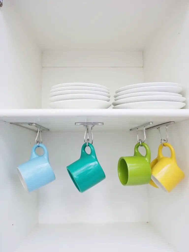 Metal under-shelf hooks installed in a kitchen cabinet, holding various colorful mugs to save space.